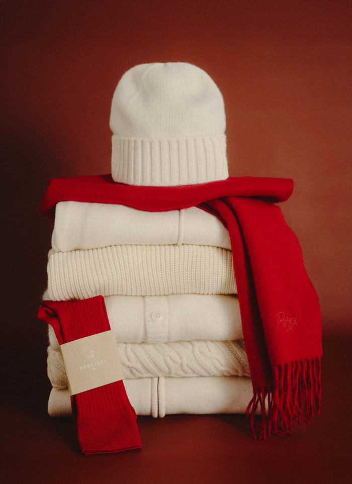 The Percival ecru wool beanie sitting on a stack of folded knitwear, styled with a festive red scarf and socks.