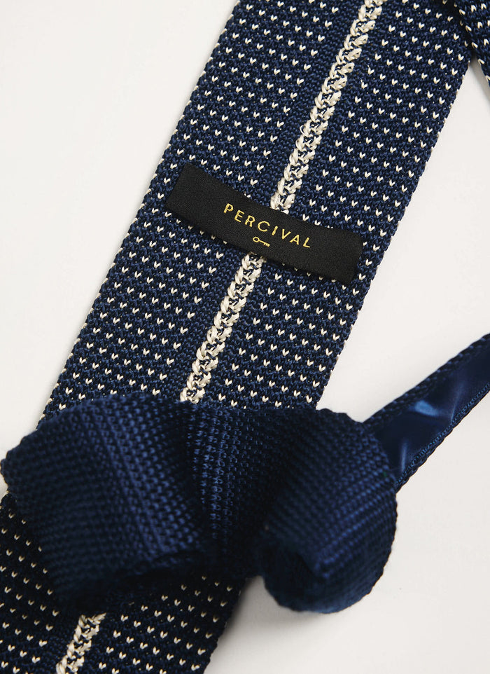 A close-up of the Percival Birdseye Silk Tie in blue, showing the intricate knitted texture and the black brand label.