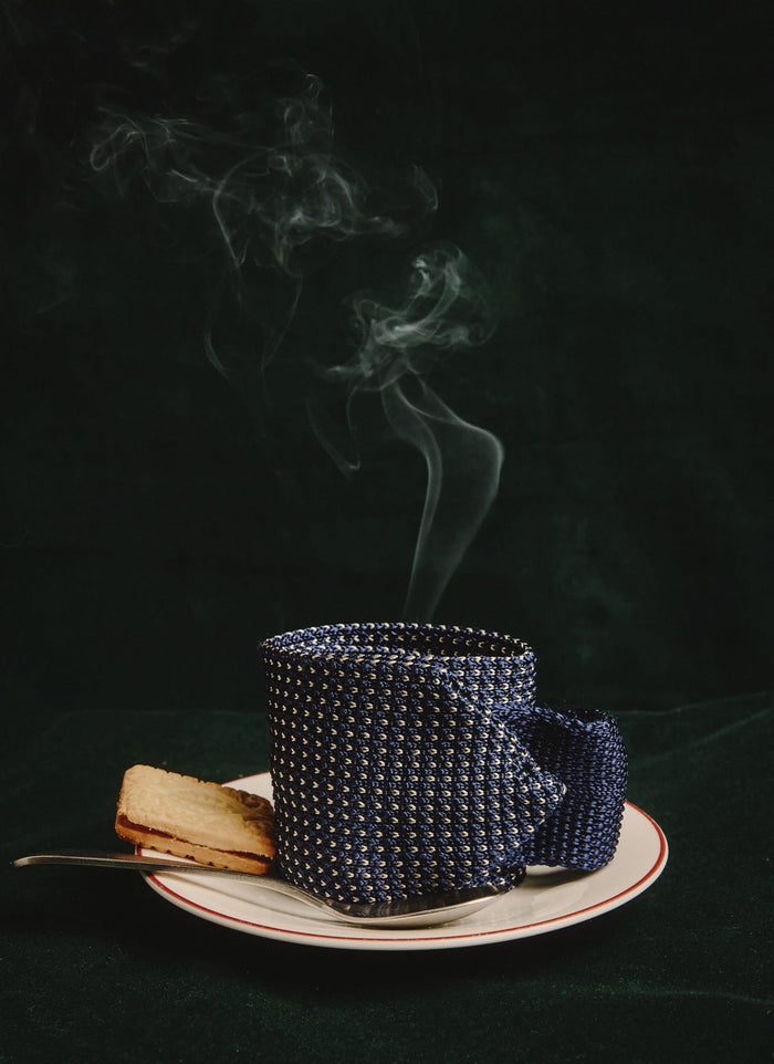 A creative still life of the Percival blue Birdseye Silk Tie, shaped into a steaming mug on a saucer with a biscuit.