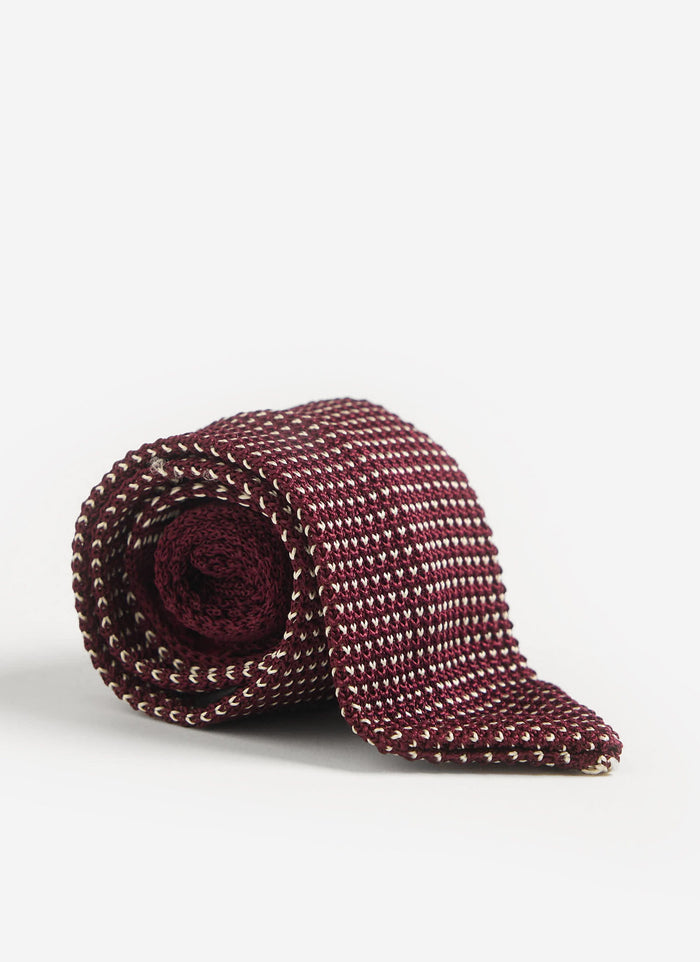 The Percival Birdseye Silk Tie in red, rolled neatly to showcase its intricate knit texture on a white studio background.