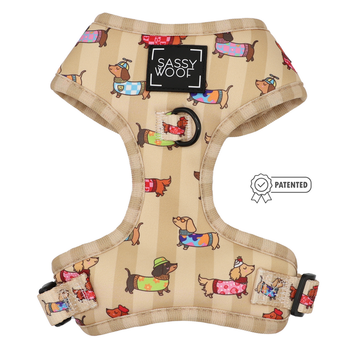 Sunbean Dog Adjustable Harness by Sassy Woof, featuring a tan design adorned with cartoon dachshunds in colorful outfits and hats. This no pull harness includes adjustable straps, a black Sassy Woof label, and a patented symbol to the right.