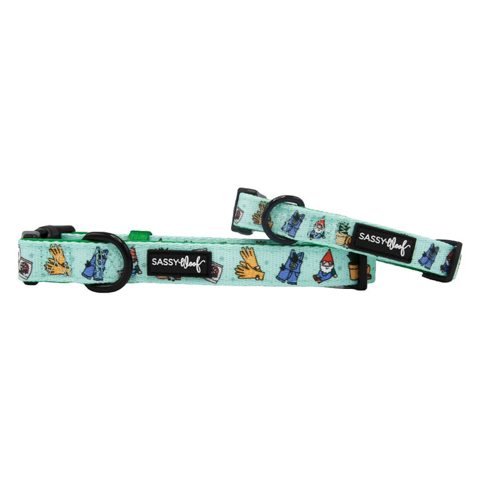 Two light blue Sassy Woof "Chillin with my Gnomies" dog collars featuring garden-themed illustrations—gloves, gnomes, watering cans—a black brand logo tag, and secure leash attachment on each.