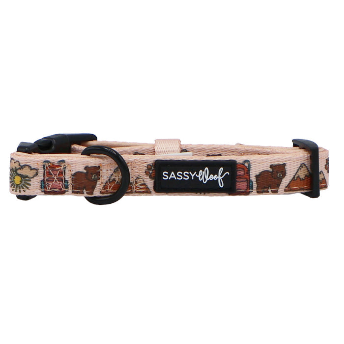 The Dog Collar - National Bark by Sassy Woof is a beige, adjustable neoprene collar with black hardware, bears, trees, and books design. A black patch reads “SASSY WOOF”—ideal for fans of outdoor dog accessories.