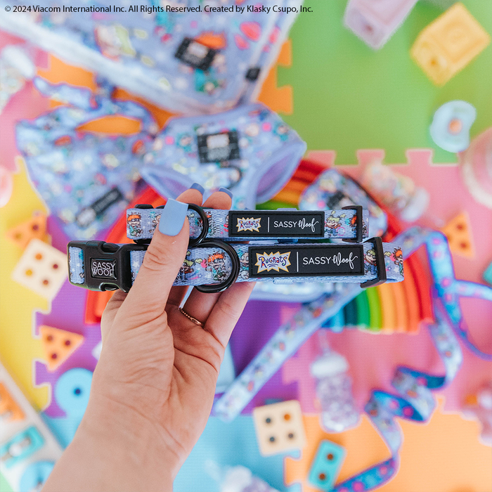 A hand with blue nail polish holds two Sassy Woof Dog Collar - Nickelodeon Rugrats™ accessories. Pastel toys and a rainbow mat in the background create a playful, vibrant 90s-inspired scene.