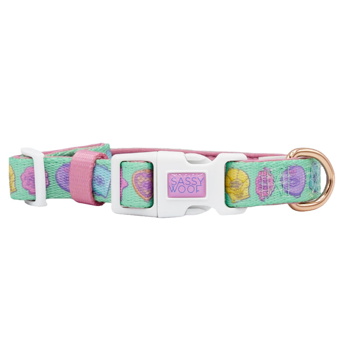 The Dog Collar - Polly Pocket™ by Sassy Woof features a pastel seashell pattern, an adjustable fit, a white plastic buckle labeled "Sassy Woof," and a gold metal D-ring for leash attachment.