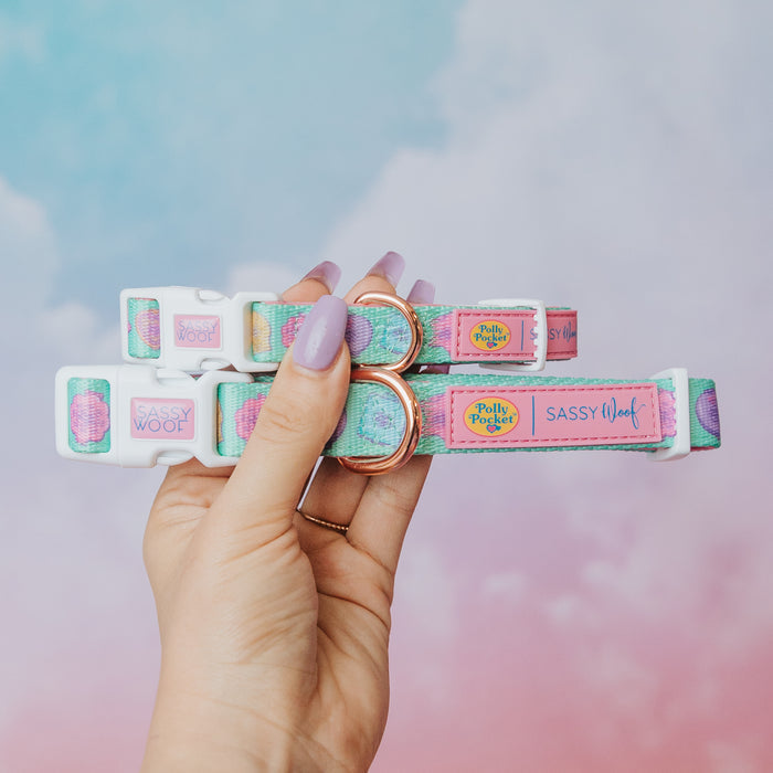 A hand with lavender nail polish displays the Sassy Woof Collar Three Piece Bundle - Polly Pocket™ against a dreamy pink and blue sky, showing off these cute pastel pet accessories with “Polly Pocket” and “Sassy Woof” labels.