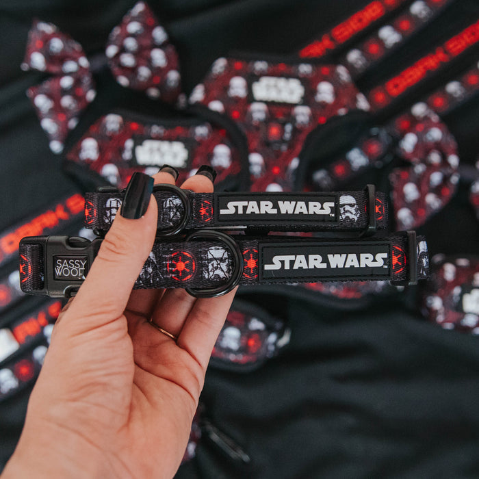 A hand holds two Sassy Woof Dog Collars – STAR WARS™ The Dark Side, each with a sturdy D-ring and featuring Star Wars logos and Stormtrooper designs. More Star Wars-themed pet accessories appear in the background.