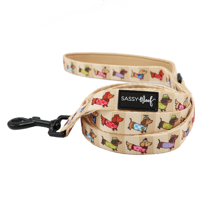Introducing the Dog Leash - Sunbean by Sassy Woof—a beige leash adorned with vibrant cartoon dachshunds sporting hats and sweaters, finished with a black clip and a signature black Sassy Woof logo patch. Perfect for pairing with your matching dog accessories!.