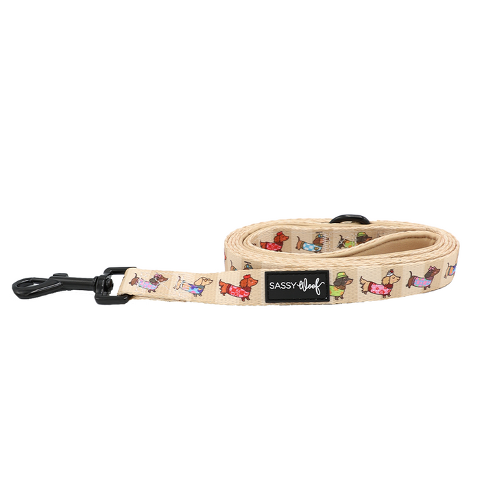 The Sassy Woof Dog Leash - Sunbean features a beige background decorated with vibrant cartoon dog illustrations and is finished with a sturdy black clasp; the stylish label reads SASSY woof—making it perfect for pairing with other matching dog accessories.