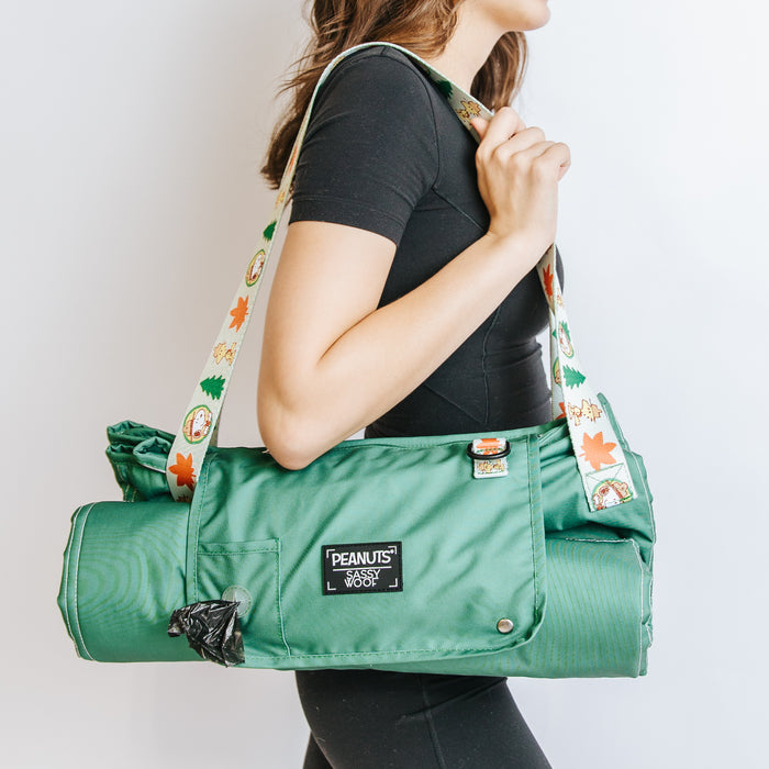 A woman in black holds the Sassy Woof Dog Picnic Blanket - Peanuts™ Beagle Scouts, a green water-resistant pet gear with a PEANUTS patch, cartoon and autumn leaf strap, and a small black item attached.