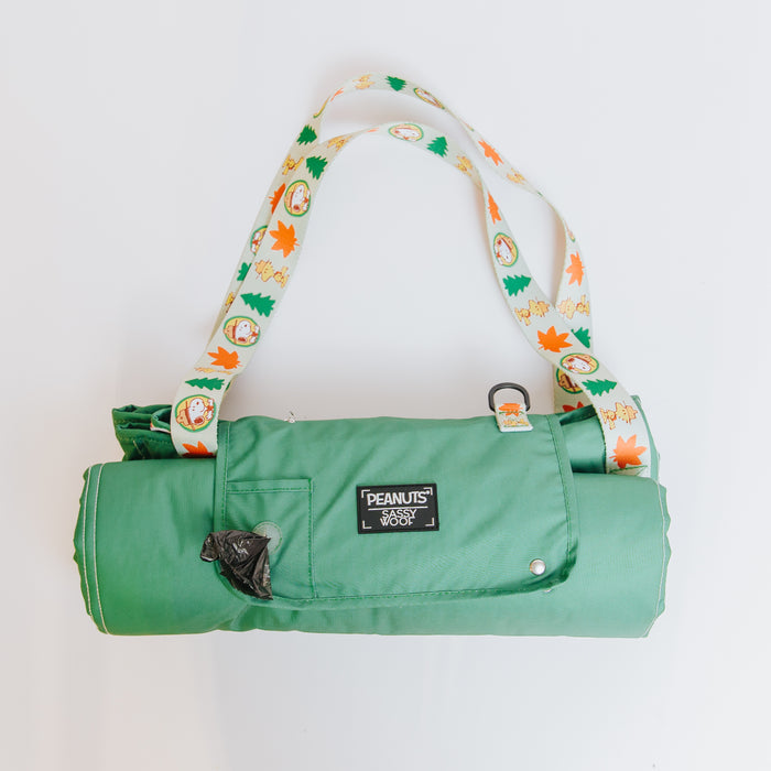 The Sassy Woof Dog Picnic Blanket - Peanuts™ Beagle Scouts is green, water-resistant, features cartoon characters and autumn leaves, a Peanuts logo patch, small black plastic clip, and is ideal for outdoor Snoopy dog accessories or pet gear.