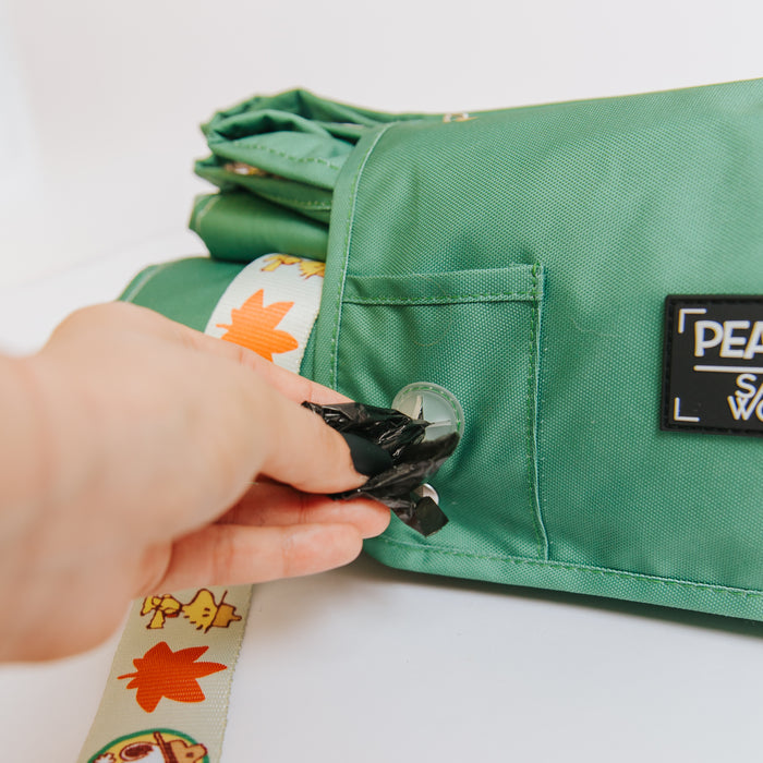 A hand pulls a black waste bag from a dispenser hole in the green, water-resistant Sassy Woof Dog Picnic Blanket - Peanuts™ Beagle Scouts, which features a decorative strap with autumn leaves and animal illustrations.