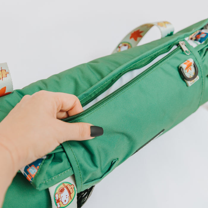 A hand with black nail polish unzips the green, water-resistant Sassy Woof Dog Picnic Blanket – Peanuts™ Beagle Scouts, featuring colorful cartoon straps, against a white background.