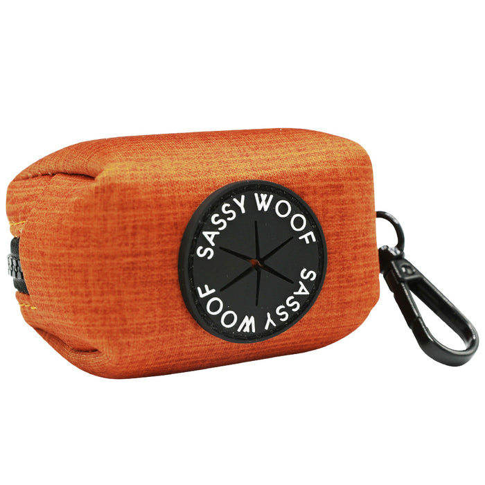 The Sassy Woof Dog Waste Bag Holder - Foxy is an orange fabric pouch featuring a black plastic clip, a round Sassy Woof patch, and a small opening for easy waste bag dispensing.