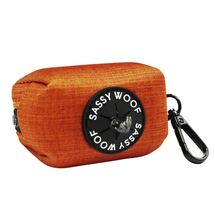 The Sassy Woof Dog Waste Bag Holder - Foxy is an orange, heavyweight neoprene holder with a black circular logo and dog image, featuring a zipper and black clip for easy leash or bag attachment.