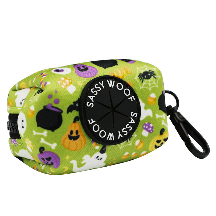 The Dog Waste Bag Holder - Haunt Mess by Sassy Woof is a green neoprene holder with Halloween designs—ghosts, pumpkins, bats—a black clip, and a front circle labeled SASSY WOOF.