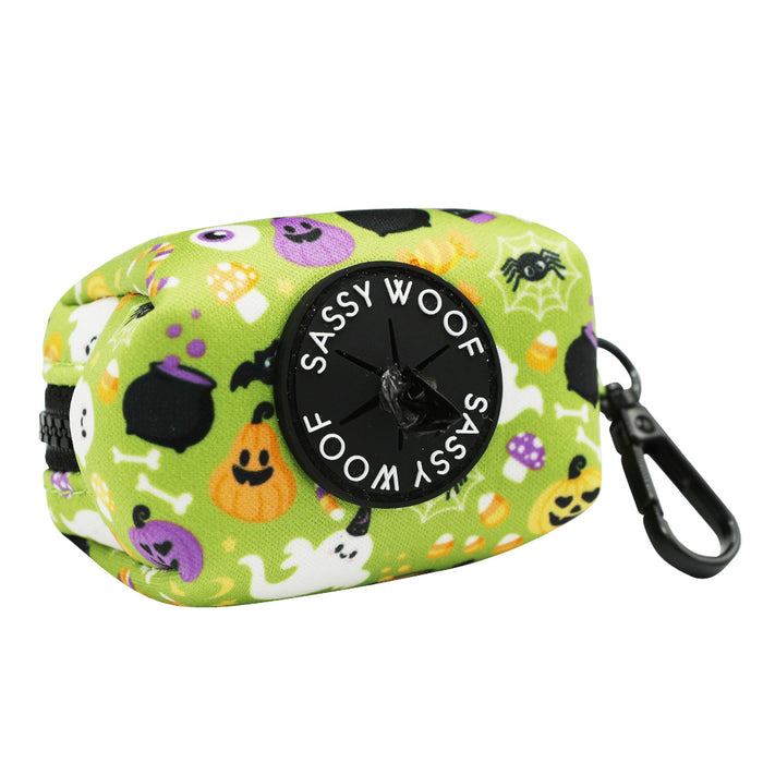 The Sassy Woof Dog Waste Bag Holder - Haunt Mess is a green neoprene holder with a Halloween pattern of ghosts, pumpkins, bats, and spiders. It features a front dispenser and black clip for easy carrying.