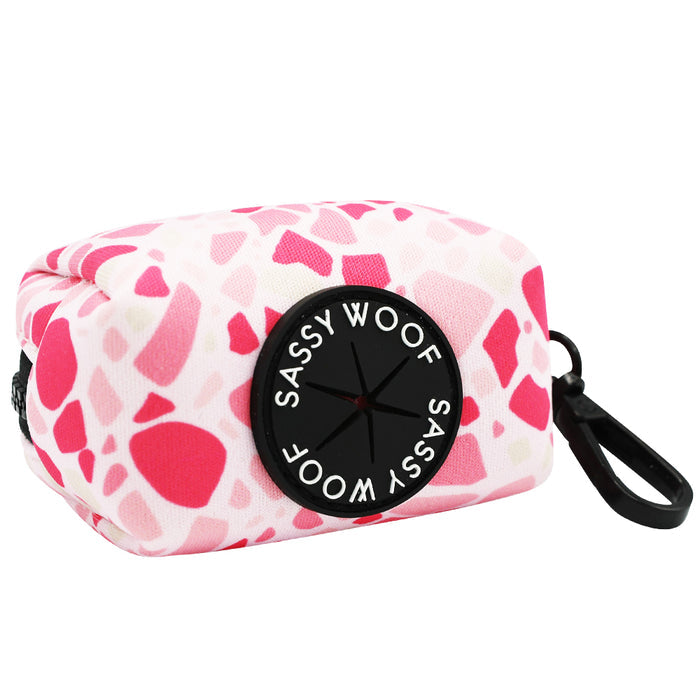The Sassy Woof Dog Waste Bag Holder – Mykonos features a rectangular pink and white pattern with a black clip and label, perfect for holding waste bags and pairing with matching dog accessories.