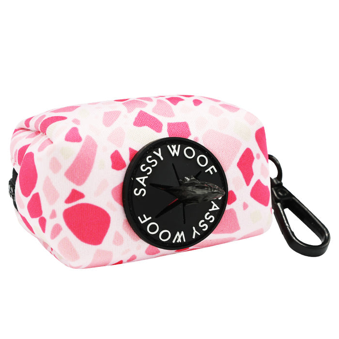 The Sassy Woof Dog Waste Bag Holder - Mykonos features a rectangular pink and white pattern, black clip, and round front label—perfect for carrying waste bags and pairing with matching dog accessories.