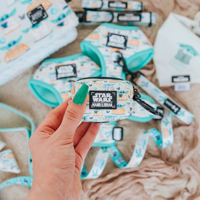 A hand with green nails holds the Sassy Woof Dog Waste Bag Holder - STAR WARS™ The Child™, a colorful Mandalorian-themed accessory. Matching Star Wars dog gear is in the background, making this holder a fun leash addition.