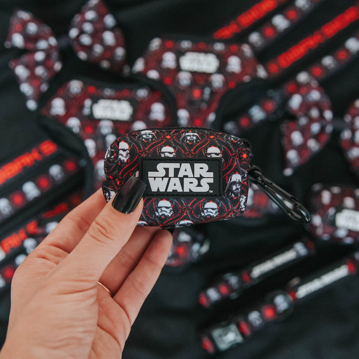 A hand with black nail polish holds the Sassy Woof Dog Waste Bag Holder - STAR WARS™ The Dark Side, featuring a stormtrooper design. Matching themed dog accessories are scattered on the dark surface in the background.