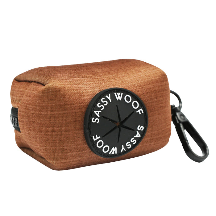 The Sassy Woof Dog Waste Bag Holder - Teddy is a brown neoprene dispenser featuring a black circular "SASSY WOOF" patch and a black clip for attaching to your leash or bag.