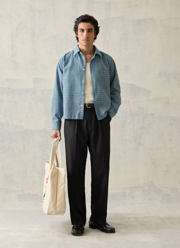 Black double pleated lyocell trousers by Percival, styled with a blue patterned shirt and a cream tote bag.