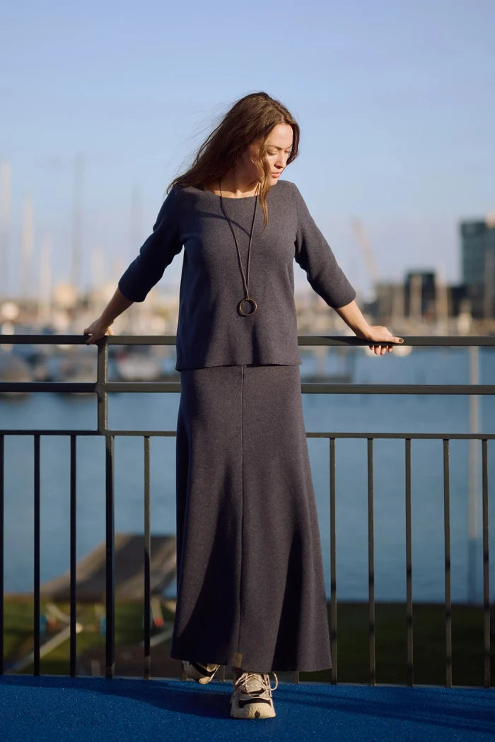 Woman in a long dark blue top and skirt standing by a waterfront with boats in the background