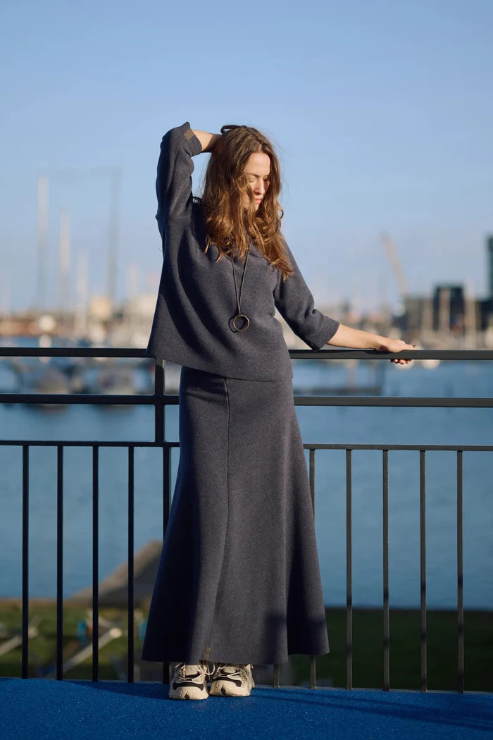 Woman in a long navy top and skirt standing by a waterfront with boats and buildings in the background.