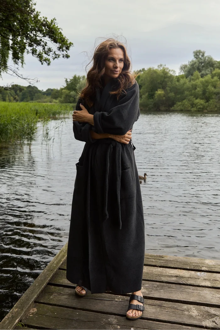 A woman wearing a long black robe stands on a wooden dock by a lake, with trees and water in the background.