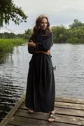 A woman wearing a long black robe stands on a wooden dock by a lake, with trees and water in the background.