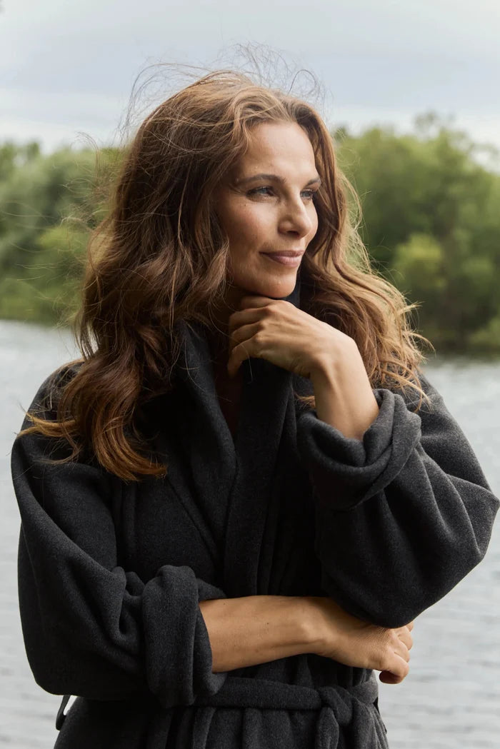 A woman with long wavy brown hair wearing a dark charcoal gray robe, posing outdoors with a lake and green trees in the background.