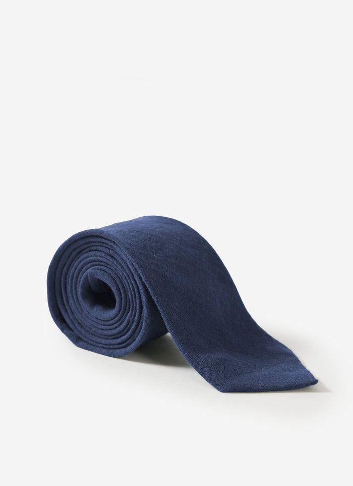 The Percival Linen Tie in navy, rolled up to display its textured fabric against a white studio background.