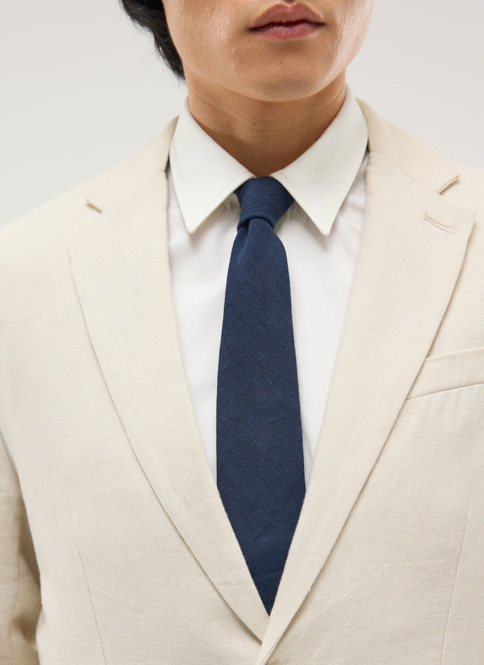 A close-up of a model wearing the Percival navy linen tie, paired with a white shirt and a cream linen suit jacket.