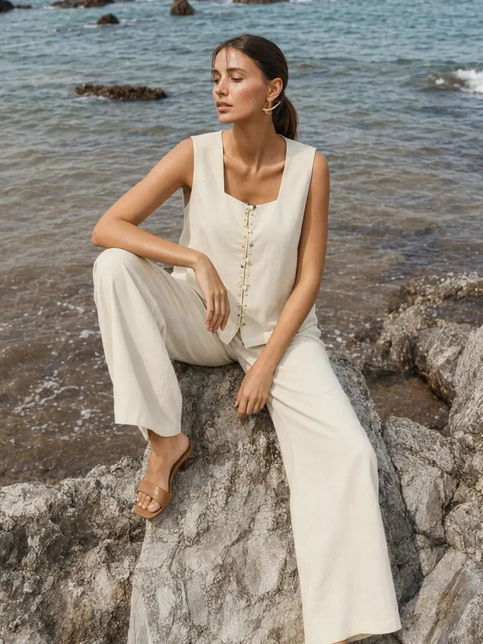 A woman sits on a rocky shoreline wearing a sleeveless cream-colored linen-like jumpsuit with wide-leg pants and tan flat sandals, with the ocean in the background.