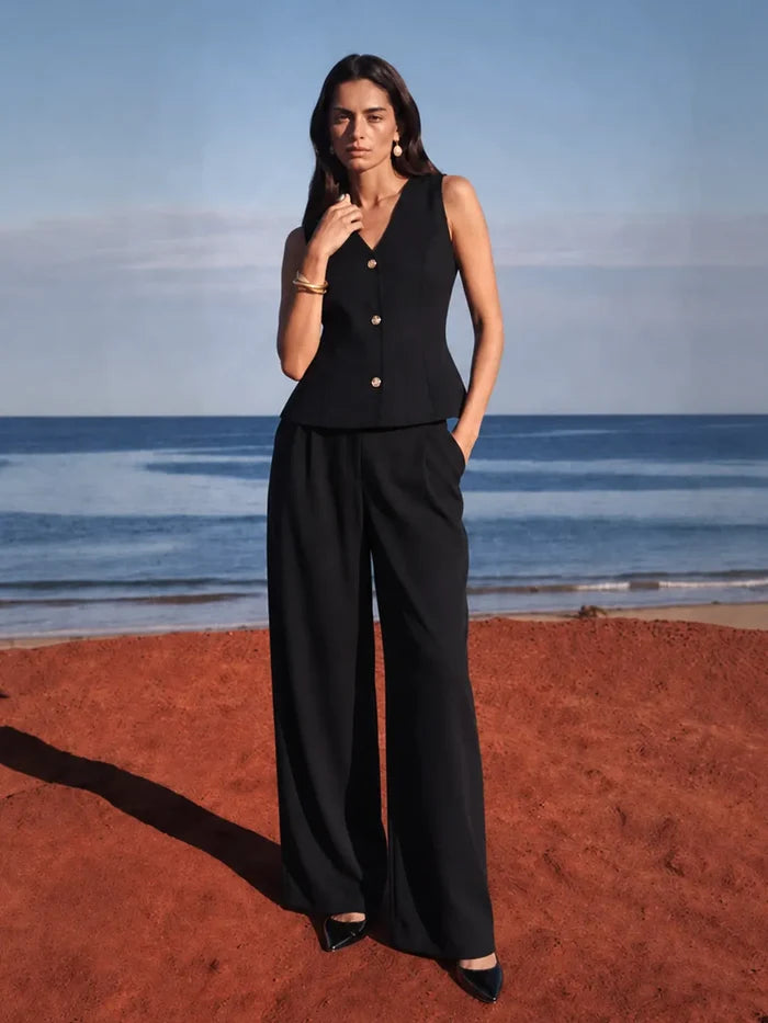A woman stands on a beach wearing a black sleeveless vest with gold buttons and matching wide-leg trousers. The background shows the blue ocean and sky, with reddish-brown sand in the foreground.