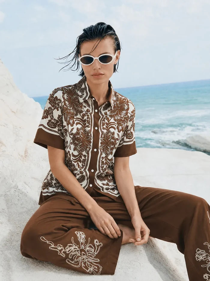 Person wearing a brown and white patterned shirt and pants sitting on a rocky beach.