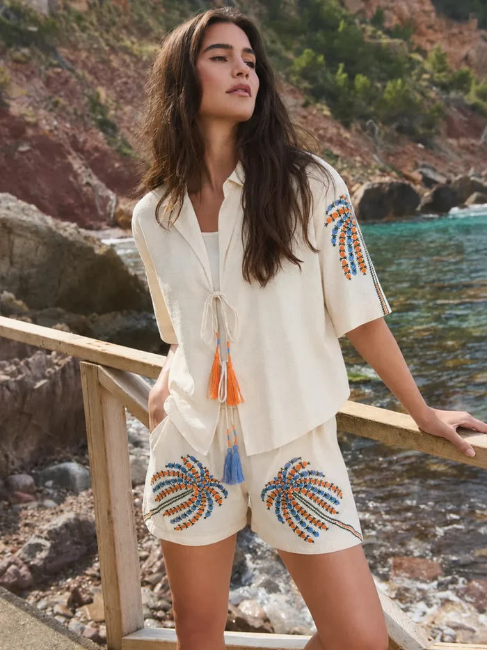 Woman in a cream outfit with decorative patterns standing by a coastal area.