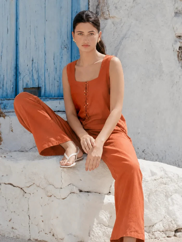 A woman wearing a rust-orange sleeveless jumpsuit with a square neckline and button-front detail, paired with matching wide-leg pants, sitting on a white stone wall in front of a blue wooden door.