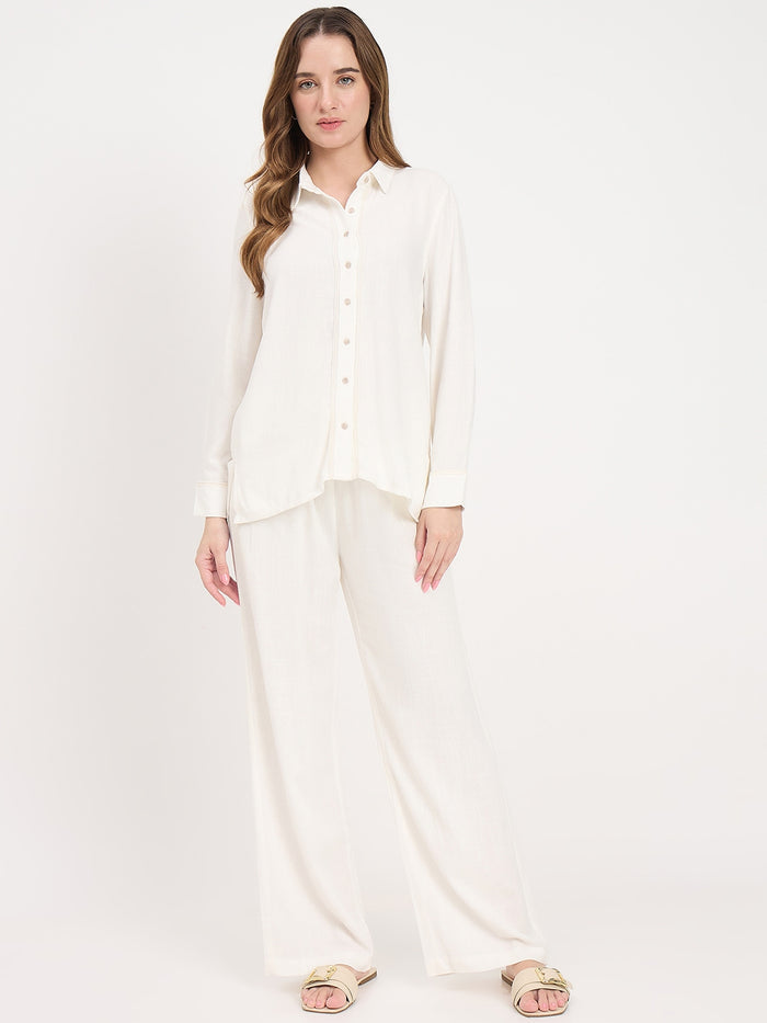 A woman modeling a matching two-piece lounge set consisting of a long-sleeve button-up shirt and wide-leg pants in a solid off-white color, paired with gold-toned slide sandals, against a plain white studio background.