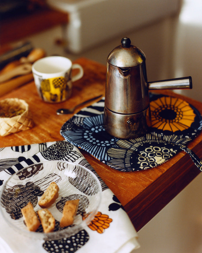 Silver espresso pot on a wooden table with a cup, cookies, and a plate of bread.