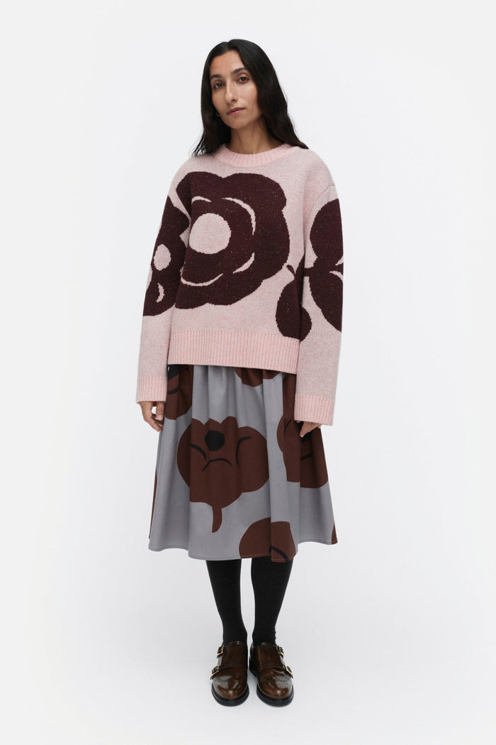 Woman wearing a pink sweater with brown floral patterns and a gray skirt on a white background