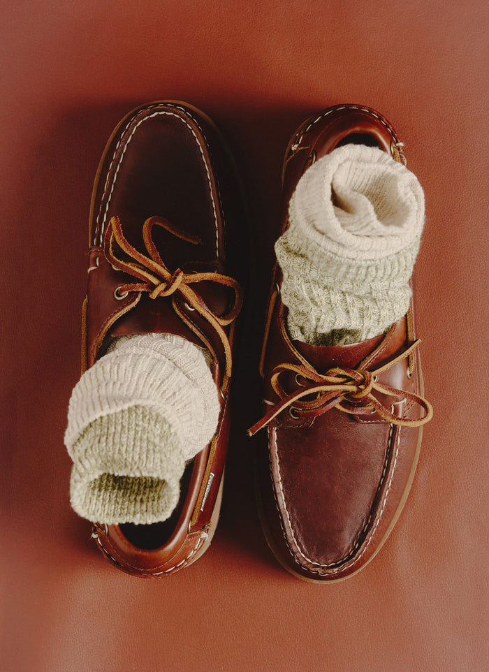 A pair of Percival's ribbed Marl Socks in oat, styled inside a pair of brown leather loafers against a tan background.