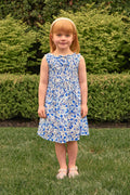 A young girl with red hair and a headband stands on green grass by trimmed bushes, wearing the Ivy City Co Mini Ana Dress in Ditsy Cobalt Floral and light-colored shoes.