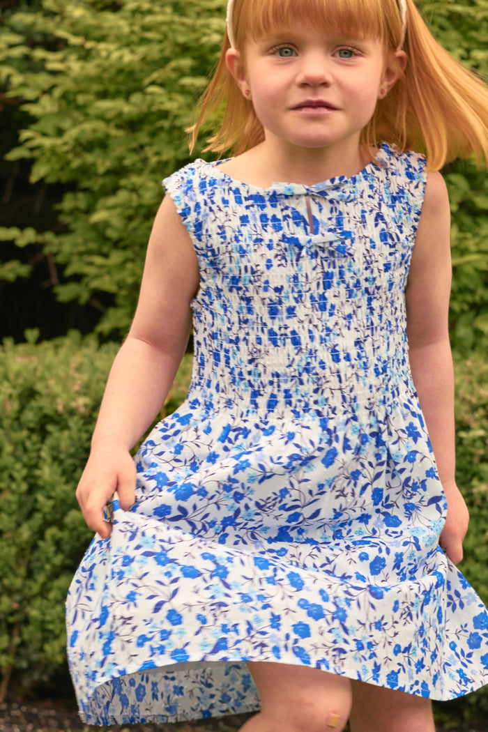 A young red-haired girl stands outdoors by green bushes, holding the edge of her Ivy City Co Mini Ana Dress in Ditsy Cobalt Floral and looking at the camera.