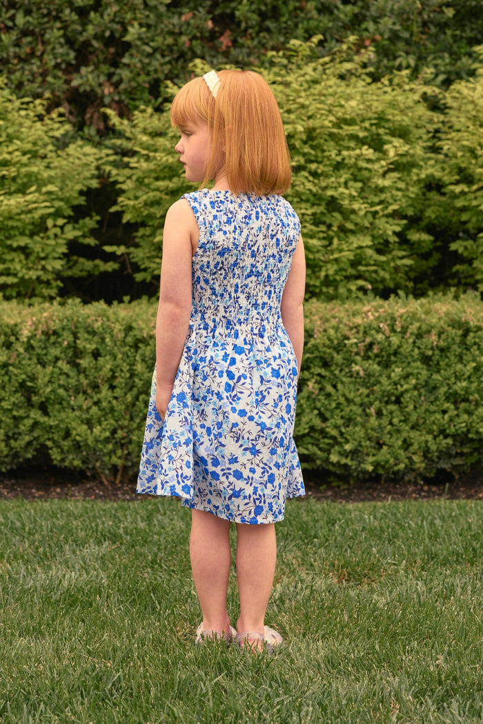 A young girl with red hair wears the Ivy City Co Mini Ana Dress in Ditsy Cobalt Floral, standing on green grass with lush bushes behind her—a charming choice for mom and me outfits.