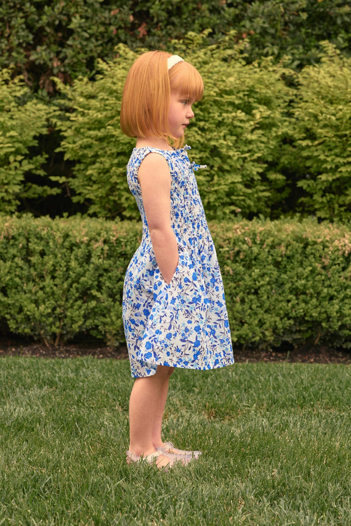 A young red-haired girl, wearing the Ivy City Co Mini Ana Dress in Ditsy Cobalt Floral and a headband, stands in profile on green grass with neatly trimmed bushes in the background.