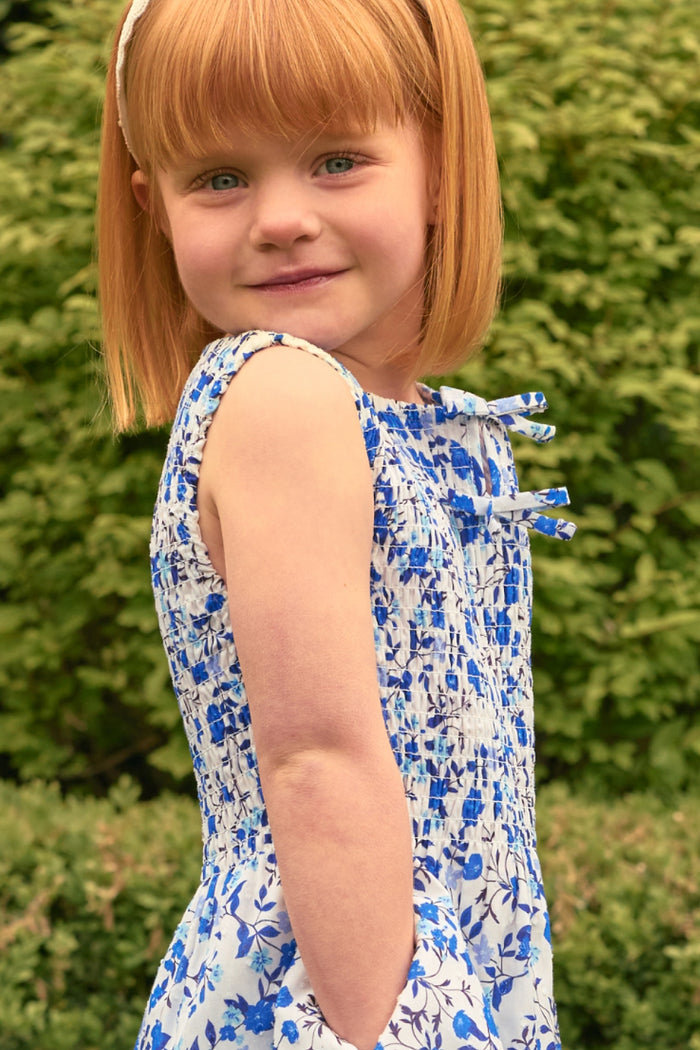 A young girl with straight red hair and blue eyes wears the Ivy City Co Mini Ana Dress in Ditsy Cobalt Floral, standing outdoors before green bushes, smiling slightly as she looks over her shoulder at the camera.