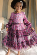 A young girl with curly hair and a white bow smiles while standing on a bed, wearing the Ivy City Co Mini Catherine Dress in Climbing Vine Bloom—a long-sleeved, ruffled floral dress perfect for matching moments.