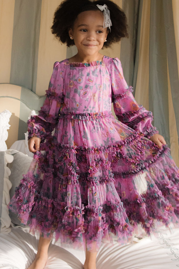 A young girl with curly hair and a white bow smiles while standing on a bed, wearing the Ivy City Co Mini Catherine Dress in Climbing Vine Bloom—a long-sleeved, ruffled floral dress perfect for matching moments.